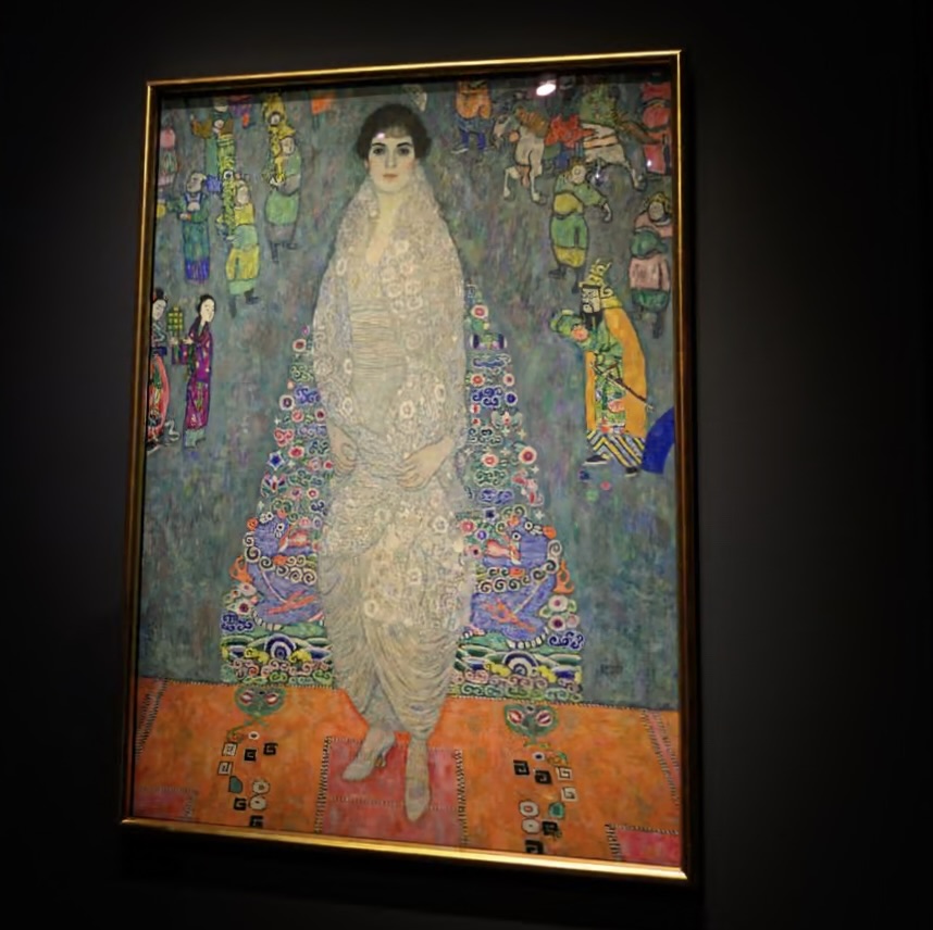 A framed full-length portrait by Gustav Klimt showing a woman in a pale, patterned gown standing against a richly detailed, colorful backdrop with stylized figures and ornamental motifs