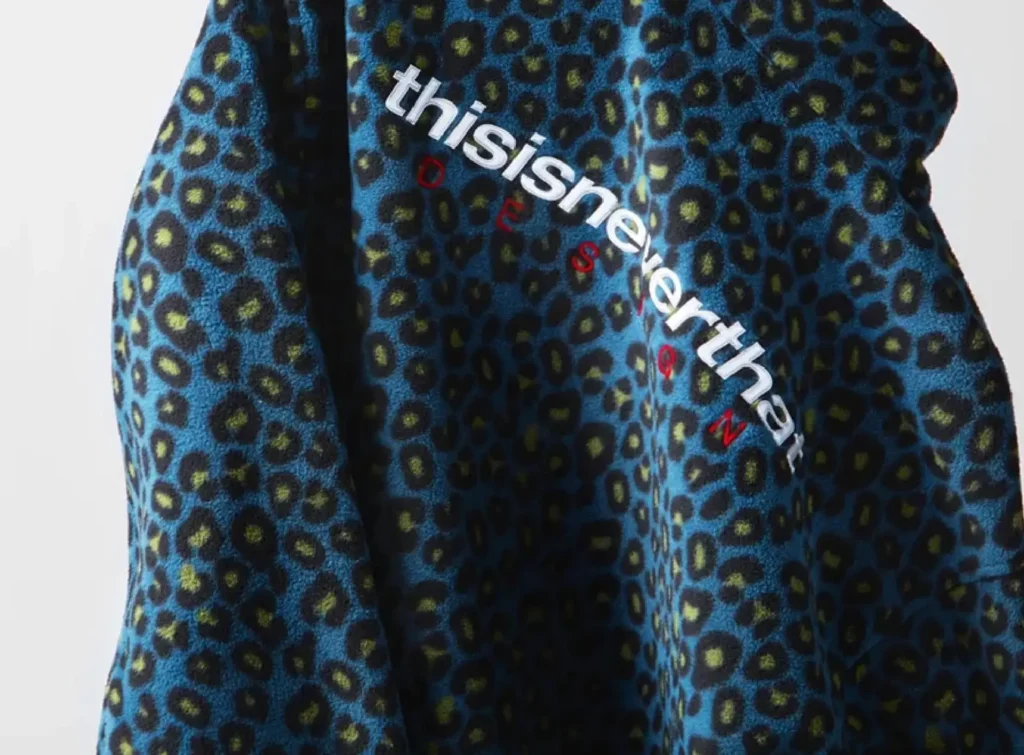 Close-up of a blue and black leopard-print fleece featuring the embroidered thisisneverthat® logo in white lettering with small red accent letters, shown draped to highlight its soft texture and pattern