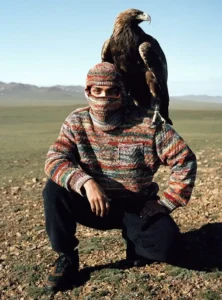 A person crouches outdoors on an open steppe landscape wearing a multicolored knit balaclava and sweater, with a large brown eagle perched on their shoulder. The scene evokes traditional falconry culture against a vast, sunlit horizon