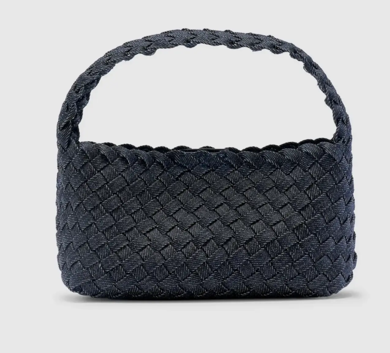 A small structured handbag woven entirely from dark indigo Japanese denim. The bag features a tightly interlaced basket-weave pattern, creating a sculptural, textured surface. It has a rounded rectangular body with softly curved edges and a matching woven top handle, giving it a cohesive, artisanal appearance. The silhouette is compact, minimal, and architectural, photographed against a clean light-grey background