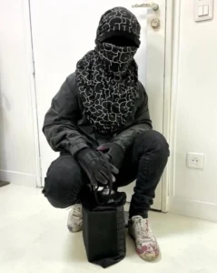 A person crouching indoors dressed in black streetwear with a patterned scarf covering their head and face, black gloves, and paint-splattered shoes, holding a black tote bag containing a spray can — a visual evocation of anonymity and graffiti culture.