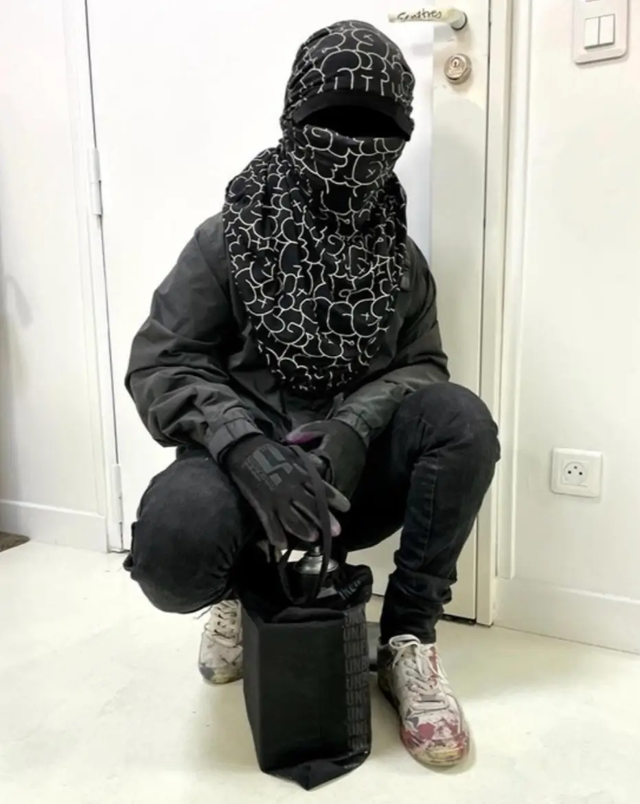 A person crouching indoors dressed in black streetwear with a patterned scarf covering their head and face, black gloves, and paint-splattered shoes, holding a black tote bag containing a spray can — a visual evocation of anonymity and graffiti culture.