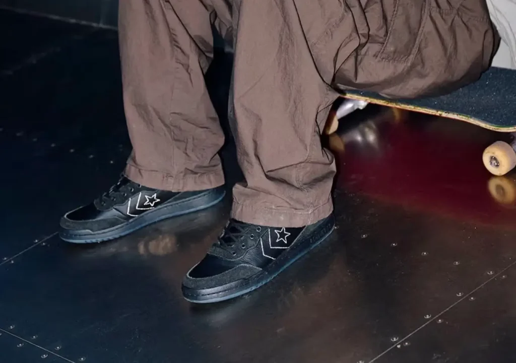 Close-up of Sage Elsesser wearing the black Converse CONS Fastbreak Pro sneakers, seated with a skateboard, showcasing the star-chevron logo and skate-ready design in a dimly lit setting