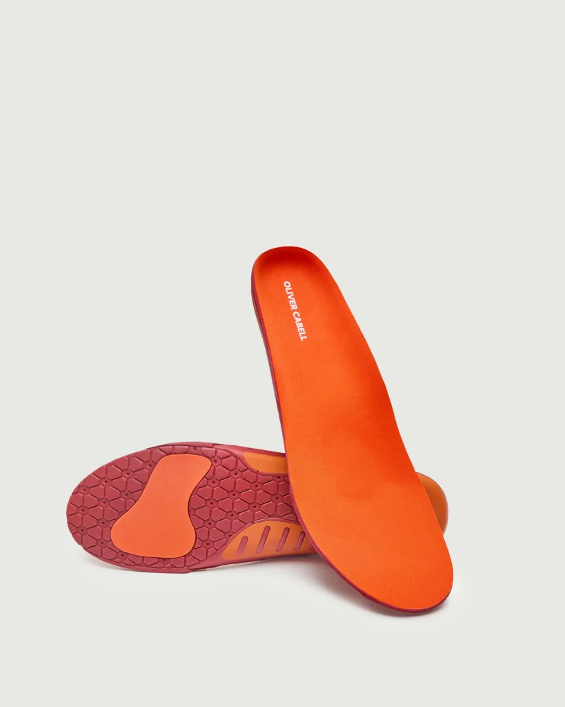 A pair of bright orange and red Oliver Cabell orthopedic insoles displayed against a light background, featuring contoured arch support, heel cushioning, and textured grip zones designed for comfort and balanced foot alignment.