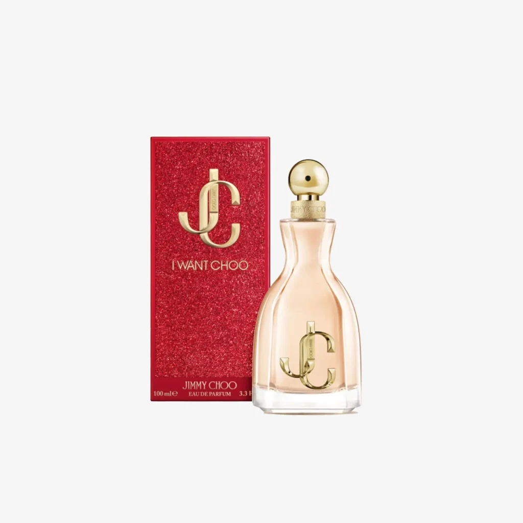 Jimmy Choo I Want Choo perfume bottle and packaging — an elegant peach-pink glass bottle with a gold spherical cap and embossed “JC” logo, displayed beside a glittering red box featuring the same gold monogram and “I Want Choo” lettering, set against a clean white background