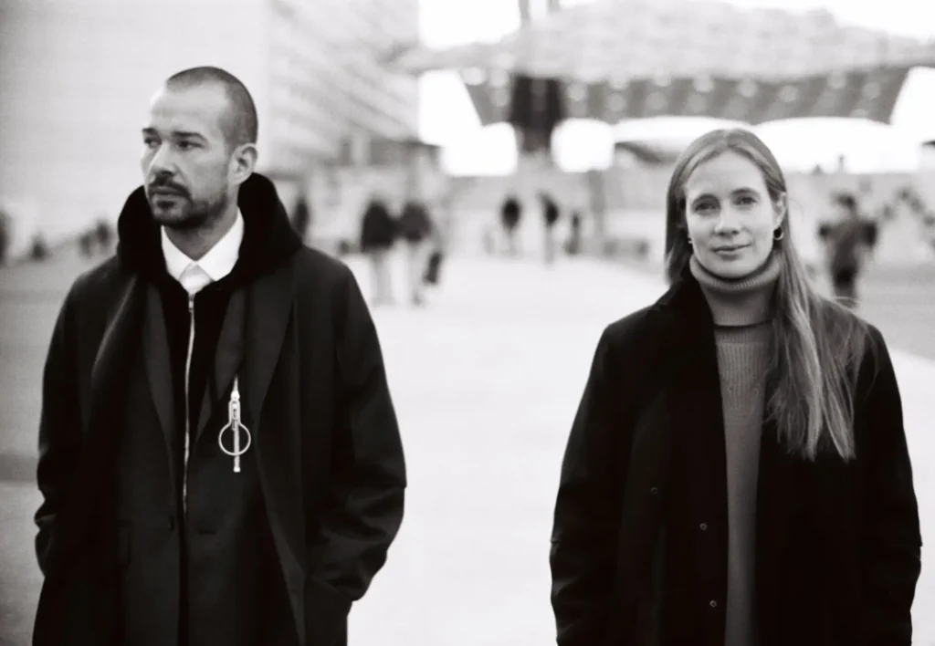 Lucie and Luke Meier Black-and-white photograph of a man and a woman standing outdoors in a modern urban plaza. Both wear dark coats; the man looks to the side with hands in his pockets, while the woman faces forward calmly. Architectural structures and blurred figures appear in the background