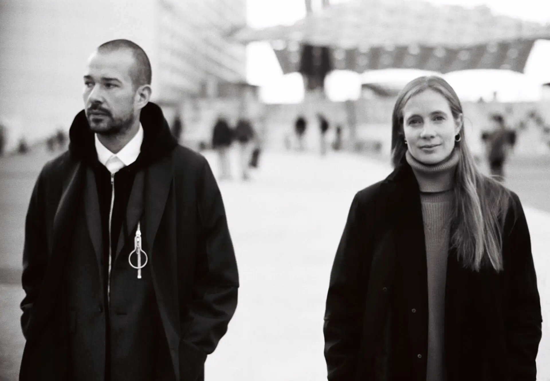 Lucie and Luke Meier Black-and-white photograph of a man and a woman standing outdoors in a modern urban plaza. Both wear dark coats; the man looks to the side with hands in his pockets, while the woman faces forward calmly. Architectural structures and blurred figures appear in the background
