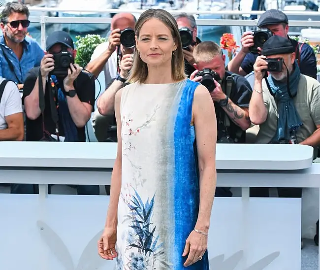 Jodie Foster standing at a Cannes photocall, wearing a sleeveless dress with a vertical blue-and-white watercolor print, while photographers in the background capture her arrival