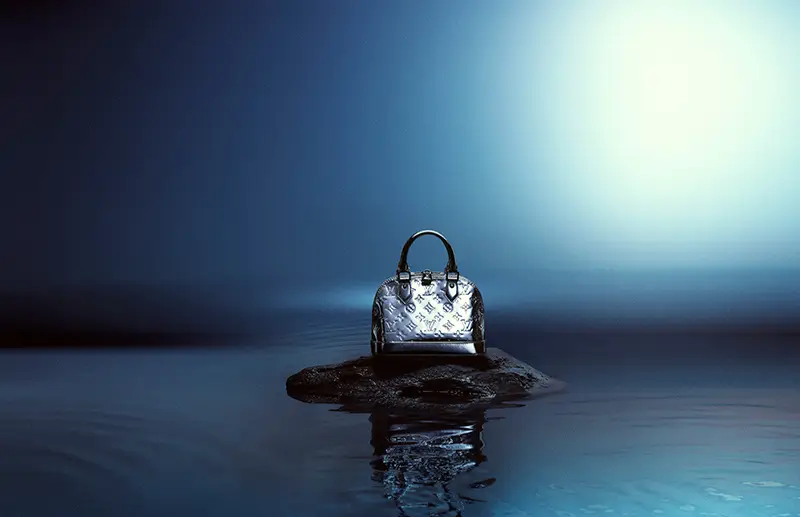 A silver Louis Vuitton Alma BB handbag sits atop a small rock surrounded by still, reflective water, illuminated by a cool, ethereal blue light that creates a serene, dreamlike atmosphere