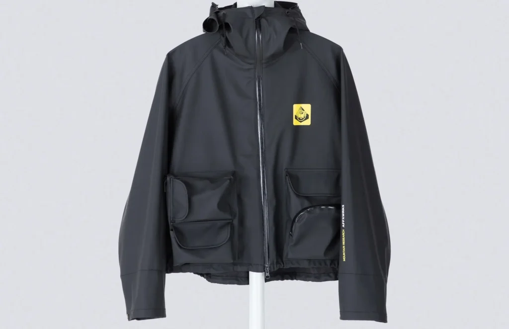 Front view of the AFFXWRKS × Mountain Research AWMR Wading Jacket in soft black, featuring a waterproof rubber-coated shell, dual front utility pockets, and a yellow co-branded patch on the chest