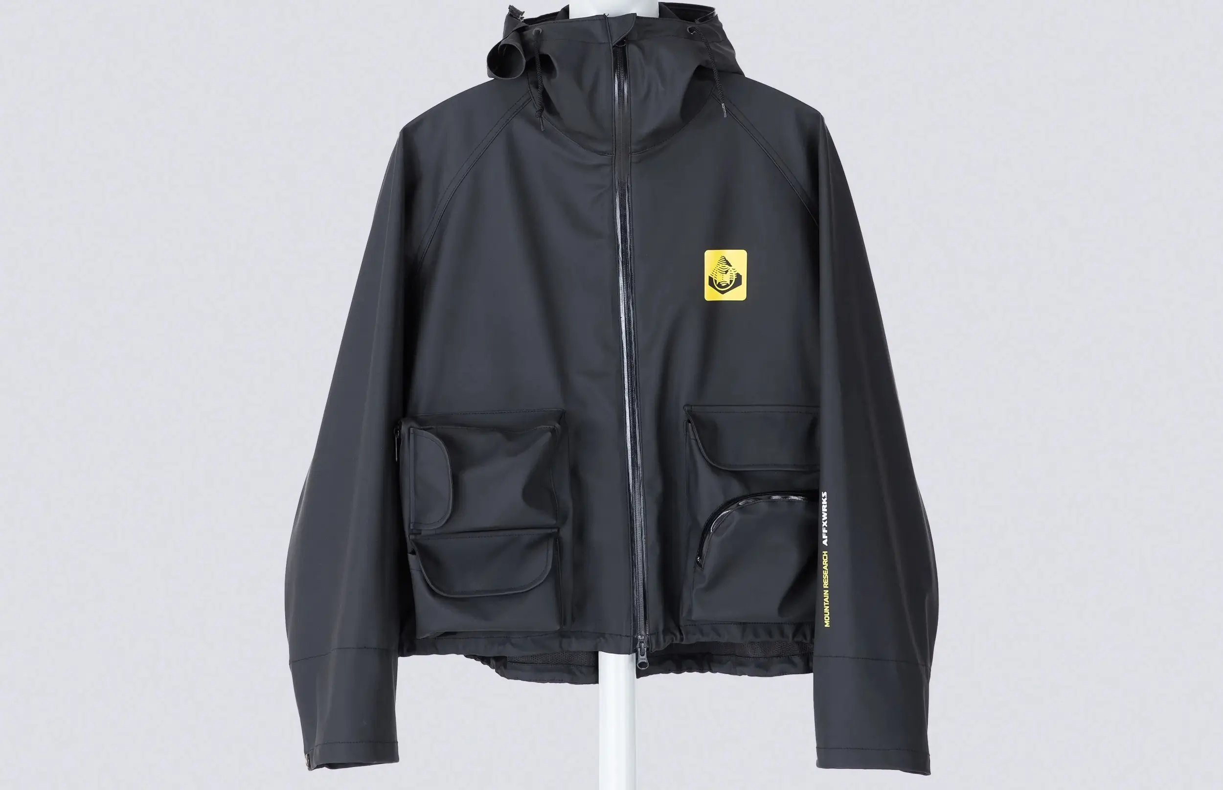 Front view of the AFFXWRKS × Mountain Research AWMR Wading Jacket in soft black, featuring a waterproof rubber-coated shell, dual front utility pockets, and a yellow co-branded patch on the chest