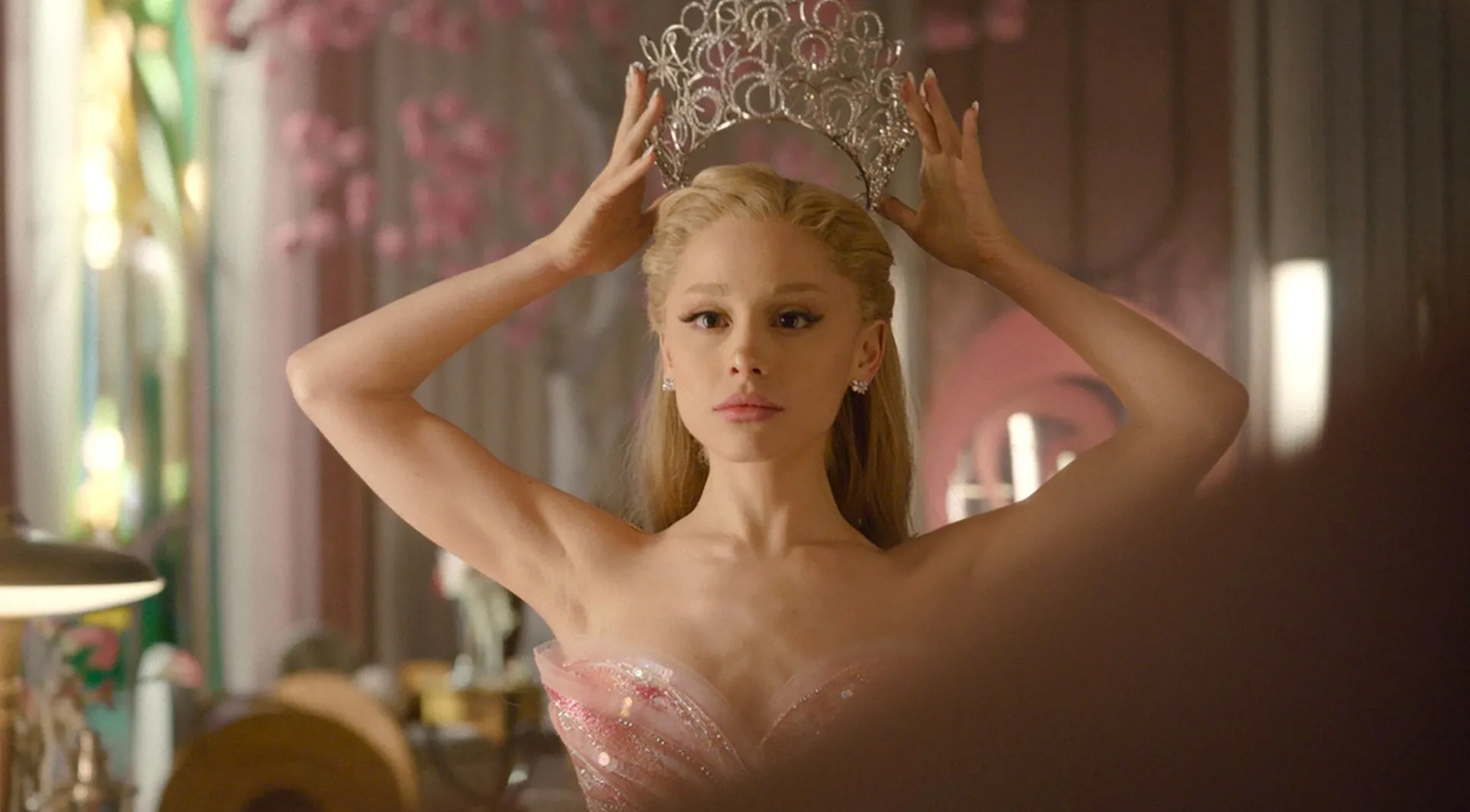 Ariana Grande dressed in a shimmering pink gown adjusts an ornate silver crown on her head while standing in front of a softly lit vanity, her expression calm and focused as warm, pastel-toned décor fills the background