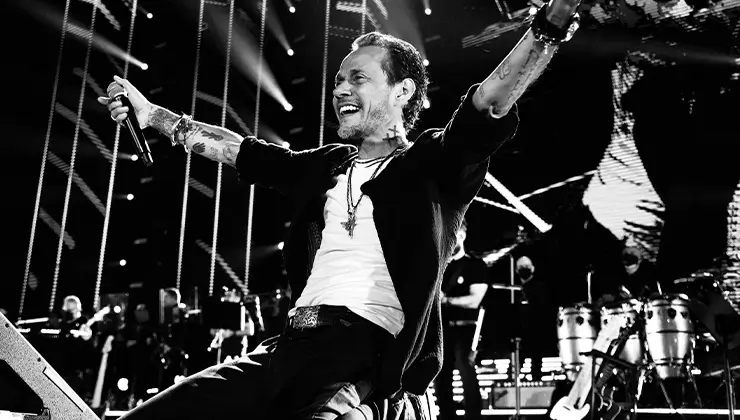 Marc Anthony performs passionately on stage during a live concert, arms outstretched in triumph, surrounded by musicians and dramatic stage lighting in grey