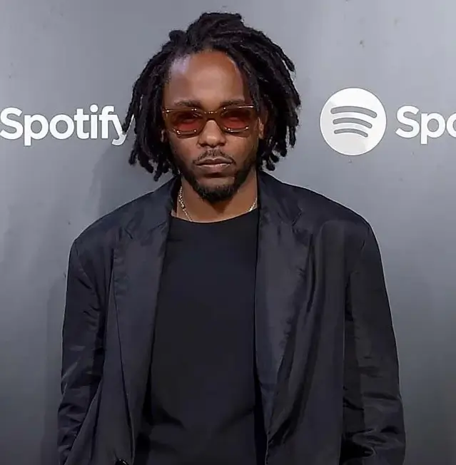 Kendrick Lamar wearing a black blazer, dark shirt, and tinted sunglasses stands in front of a grey Spotify-branded backdrop, posing confidently for event photos