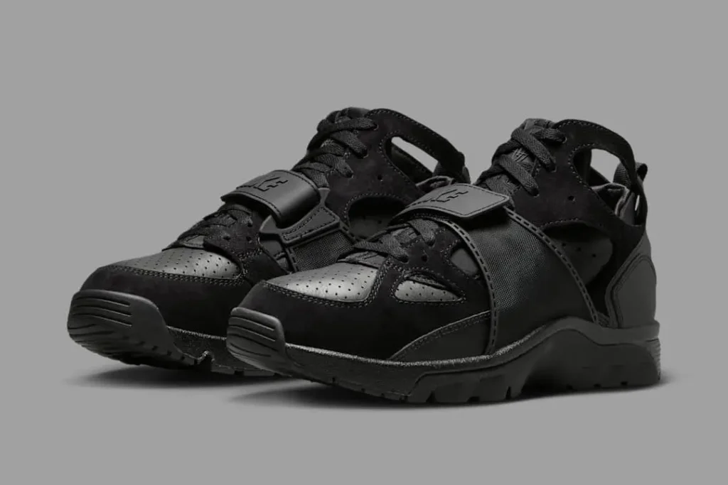 Nike Air Trainer Huarache “Triple Black” sneakers — a monochrome all-black pair featuring perforated leather panels, neoprene inner bootie, and a midfoot strap with embossed Huarache logo, displayed on a neutral gray background to highlight the shoe’s stealth design and sculpted silhouette