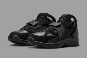 Nike Air Trainer Huarache “Triple Black” sneakers — a monochrome all-black pair featuring perforated leather panels, neoprene inner bootie, and a midfoot strap with embossed Huarache logo, displayed on a neutral gray background to highlight the shoe’s stealth design and sculpted silhouette