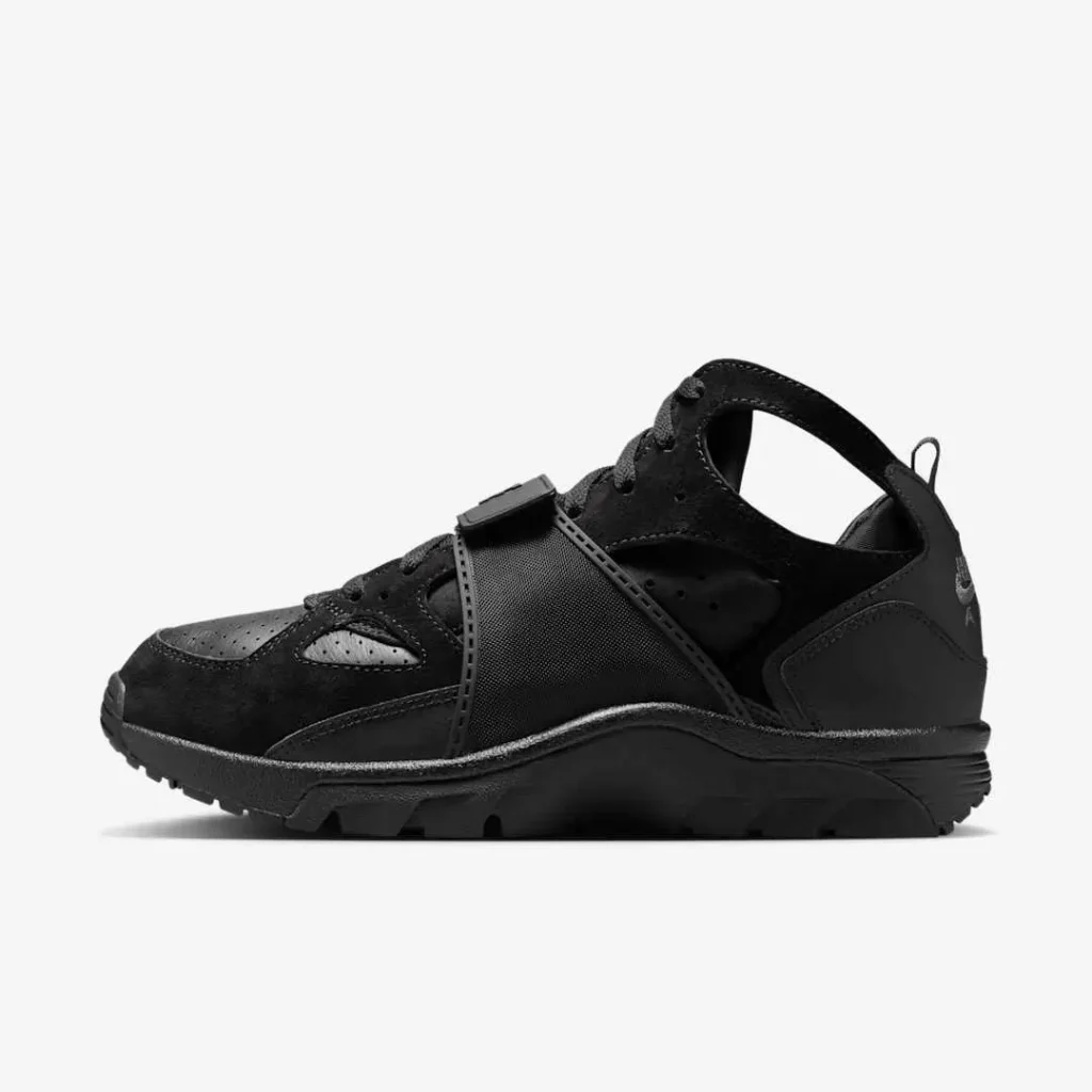 Side profile of the Nike Air Trainer Huarache “Triple Black” sneaker showcasing its matte leather and neoprene upper, adjustable midfoot strap, and cut-out ankle design, displayed against a clean white background highlighting its stealth all-black construction and sculpted midsole