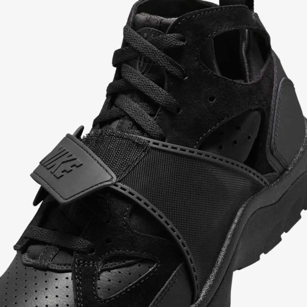 Close-up view of the Nike Air Trainer Huarache “Triple Black” sneaker, focusing on the textured midfoot strap with embossed Nike logo, black laces, and perforated leather and neoprene upper, highlighting the shoe’s layered construction and tonal detailing