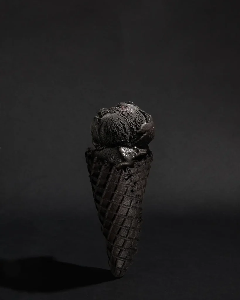 A jet-black ice cream cone photographed against a dark background, featuring two scoops of deep matte black ice cream with a matching waffle cone, highlighting smooth texture and minimal lighting for a monochrome, sculptural look