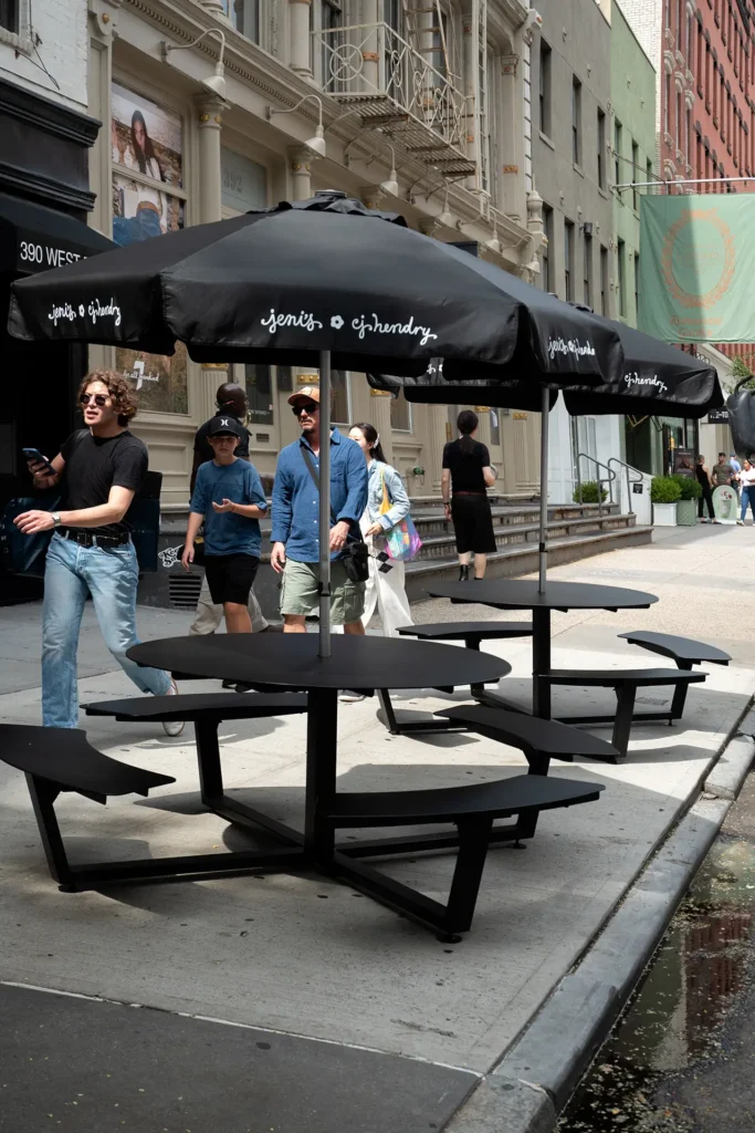 Street view of black outdoor tables and umbrellas branded with “Jeni’s x CJ Hendry,” set along a New York sidewalk as pedestrians walk by, showcasing the minimalist and modern aesthetic of the connection's pop-up setup