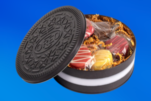 Open OREO Thanksgiving Dinner Cookie Tin shaped like a giant black OREO cookie, partially revealing six coated sandwich cookies in festive autumn colors—red, yellow, and brown—nestled in golden paper shreds against a bright blue background