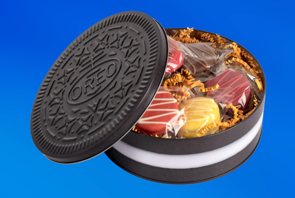 Open OREO Thanksgiving Dinner Cookie Tin shaped like a giant black OREO cookie, partially revealing six coated sandwich cookies in festive autumn colors—red, yellow, and brown—nestled in golden paper shreds against a bright blue background