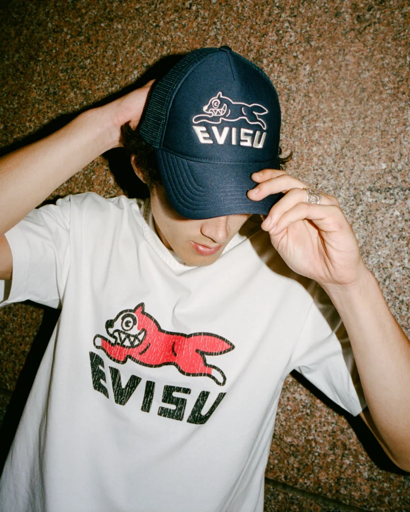 model wearing a navy Evisu x Icecream trucker cap embroidered with the Running Dog logo and matching white T-shirt featuring a red Running Dog graphic and bold EVISU lettering