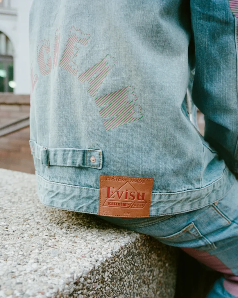 close-up of the Evisu x Icecream denim jacket showing dual branding — the pink-and-green striped ICECREAM lettering on the back and the brown leather “Custom Made Evisu Icecream & Co” patch above matching denim pants