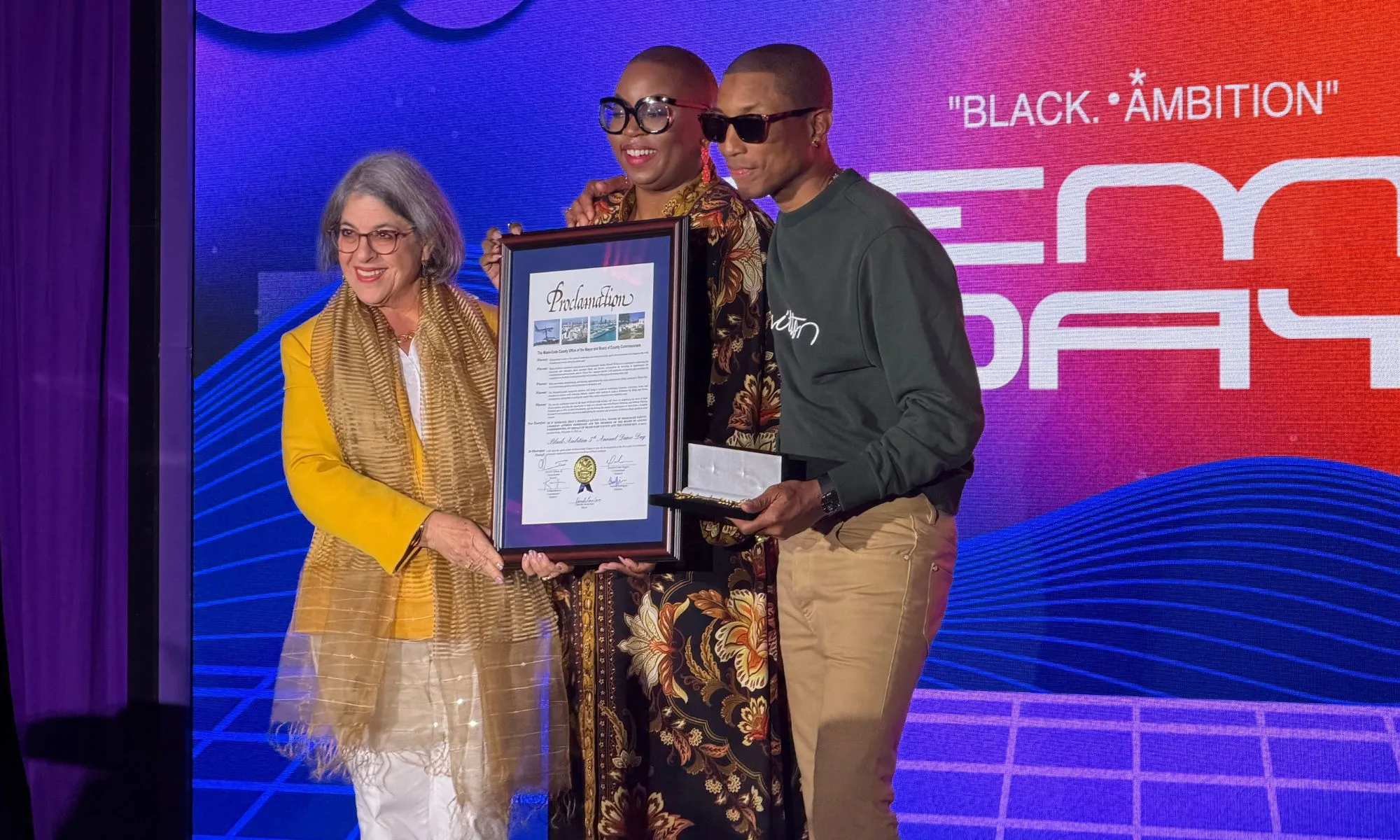 Three people stand together onstage during a Black Ambition event, holding a framed proclamation and an open presentation box. On the left, a woman in a yellow jacket and gold scarf smiles as she helps display the framed document. At the center, another person in patterned clothing holds the frame as well, while on the right, Pharrell Williams—wearing sunglasses, a dark green sweatshirt, and tan pants—holds the presentation box. A colorful digital backdrop with the words “BLACK AMBITION” and graphic wave patterns fills the background