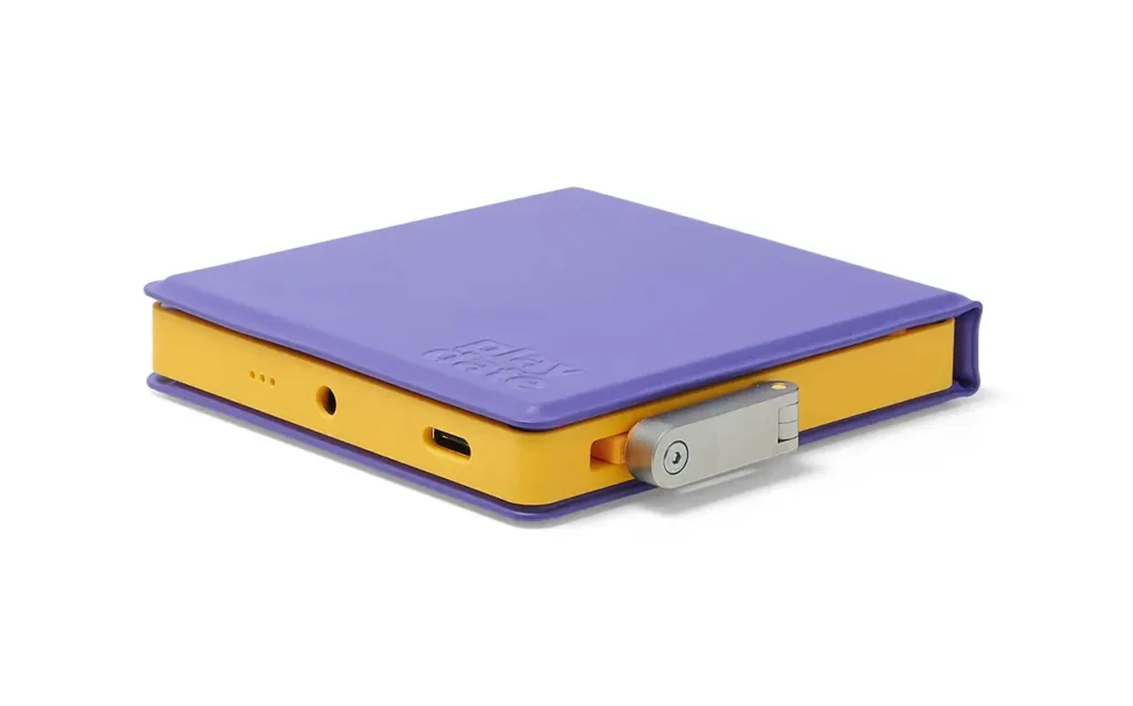 A yellow Playdate handheld gaming console partially enclosed in a fitted purple protective case, shown from a low side angle. The case covers the top and sides while leaving the metal crank, USB-C port, and headphone jack visible, highlighting the device’s compact, colorful, and playful design