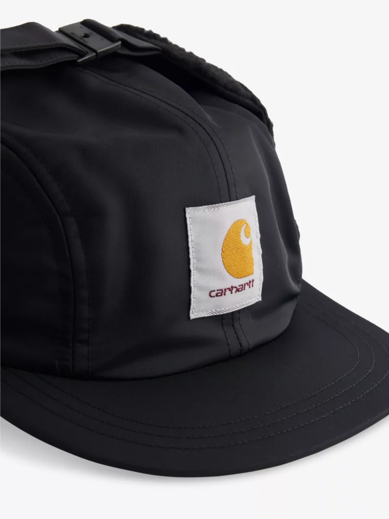 Close-up of the Carhartt WIP Oltera Ear-Flap Woven Cap in black, showing the front panel with the signature brown Carhartt square logo patch and detailed stitching on the brim