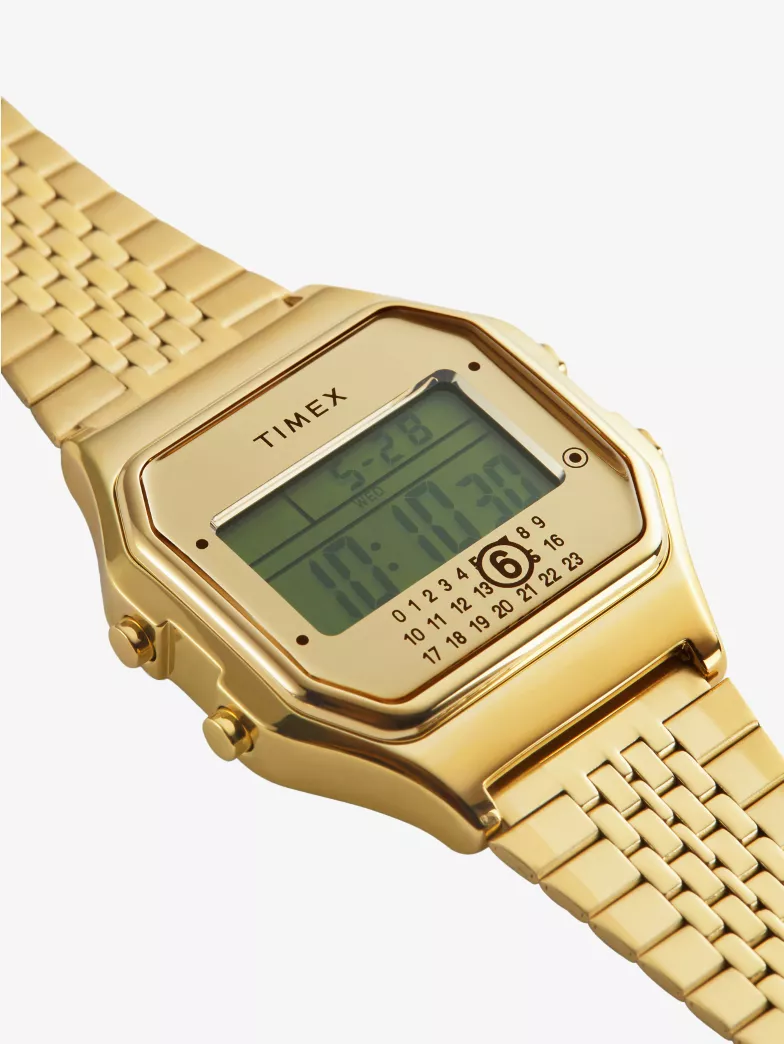 Close-up of the Timex x MM6 Maison Margiela T80 watch in gold-tone stainless steel, featuring a rectangular digital display, mirrored dial mask, and the signature MM6 numbered ring with the number 6 circled above a woven metal bracelet