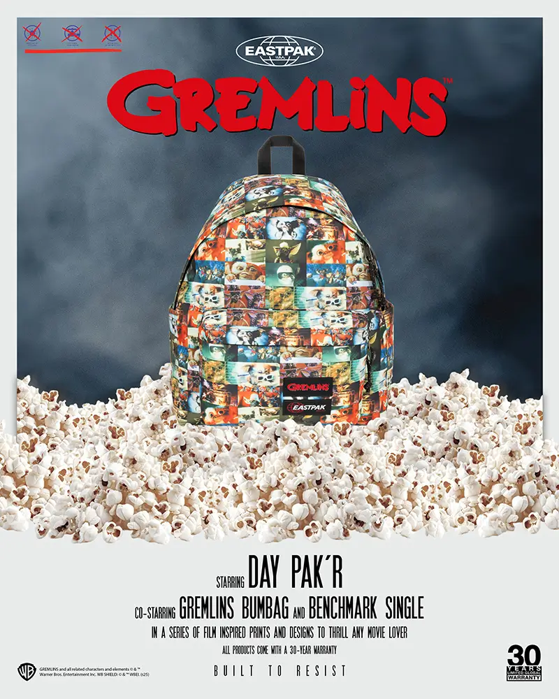 Eastpak x Gremlins promotional poster featuring a Day Pak’r backpack covered in a collage of Gizmo and Gremlins scenes, displayed on a large pile of popcorn with a smoky blue backdrop. The Gremlins logo appears in bold red above the bag, alongside film-style credits highlighting the Day Pak’r, Gremlins Bumbag, and Benchmark Single, with Eastpak and 30-year warranty branding