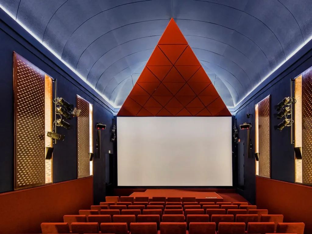 Interior view of the newly redesigned auditorium at Museo Reina Sofía in Madrid, featuring deep blue acoustic walls, rust-red seating, and a striking suspended triangular ceiling element above the projection screen that conceals the central speaker, combining geometric precision with a contemporary cinematic atmosphere