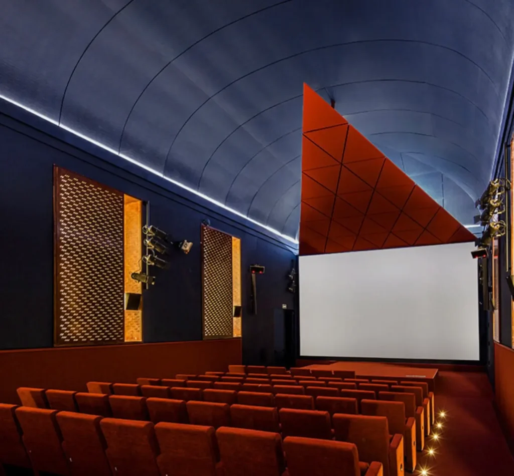 Angled view of the contemporary cinema hall at Museo Reina Sofía, showcasing red upholstered seats, perforated bronze acoustic panels, and the signature suspended triangular ceiling structure in terracotta tones beneath a softly lit arched blue ceiling