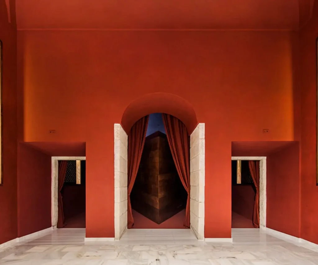 Entrance hall of the redesigned Reina Sofía auditorium featuring deep terracotta walls, marble flooring, and arched stone-framed doorways with heavy red curtains leading into the cinema, creating a dramatic transition between classical architecture and contemporary design