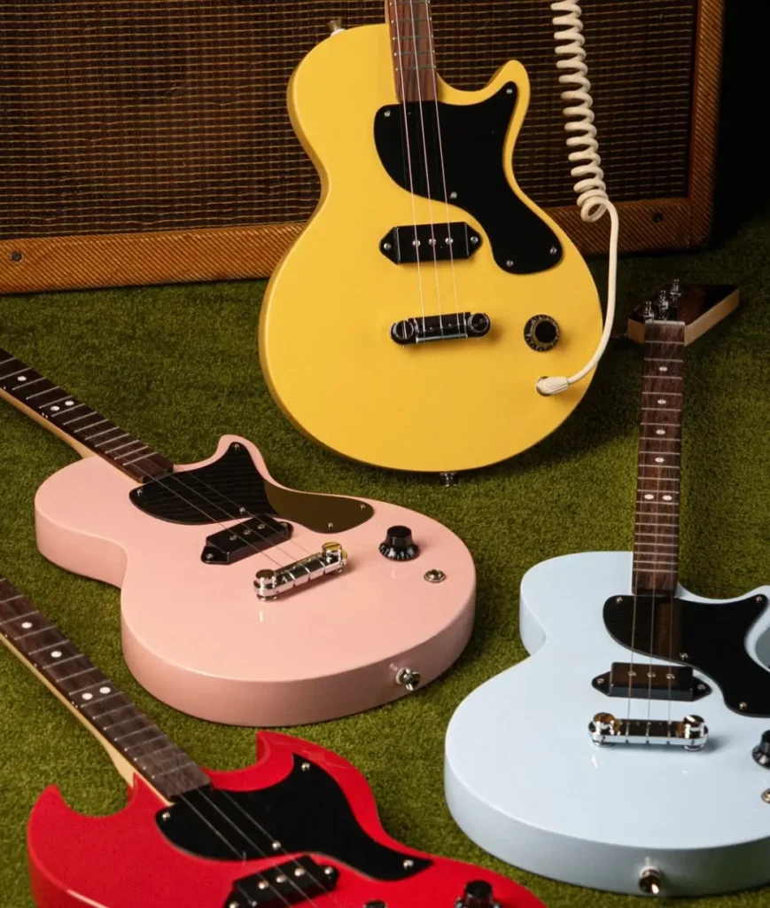 close-up of Gibson x Loog electric guitars in four colors — TV Yellow, pastel pink, light blue, and cherry red — arranged on green carpet in front of a vintage amplifier, highlighting their three-string design and compact Les Paul-inspired shape