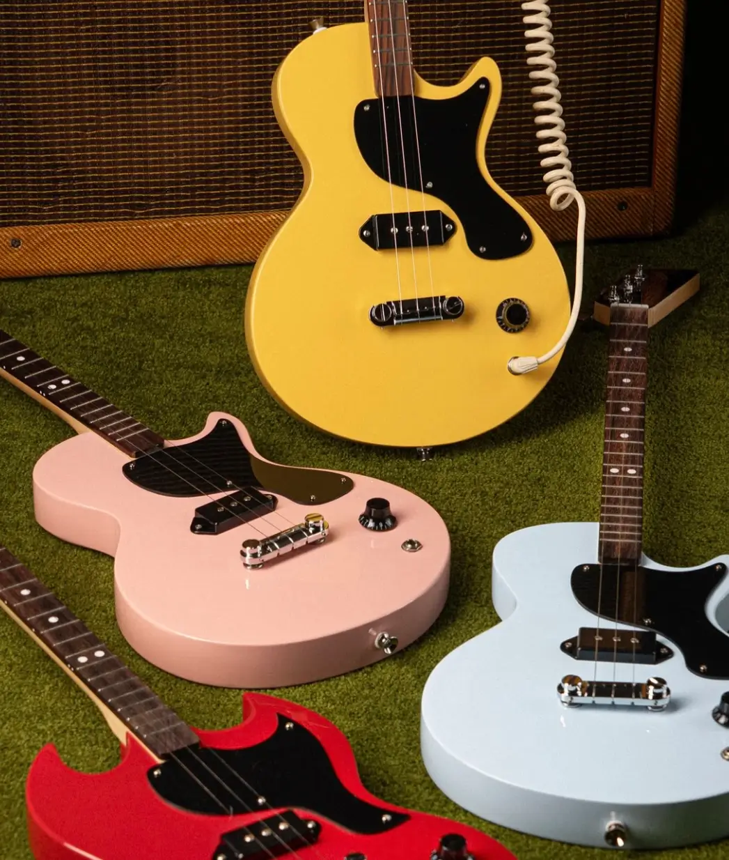 close-up of Gibson x Loog electric guitars in four colors — TV Yellow, pastel pink, light blue, and cherry red — arranged on green carpet in front of a vintage amplifier, highlighting their three-string design and compact Les Paul-inspired shape