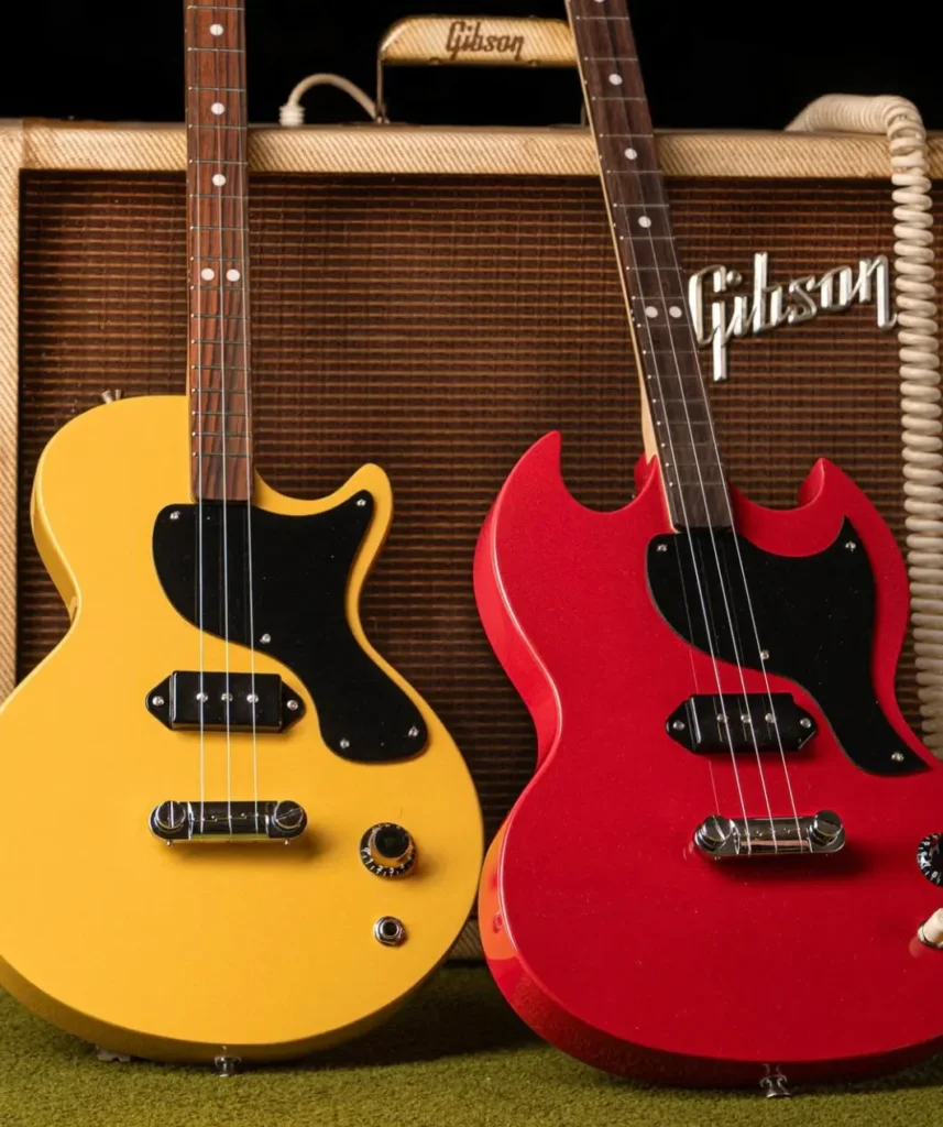 Gibson x Loog electric guitars in TV Yellow and Cherry Red finishes leaning against a vintage Gibson amplifier with coiled cable, showcasing their compact three-string design and black pickguards, blending classic Gibson style with playful Loog proportions