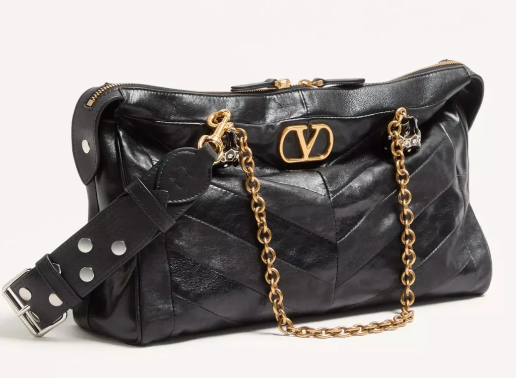Close-up of the Valentino Garavani Panthea Shoulder Bag crafted in black nappa leather with a chevron pattern. The bag features antique gold-tone chain handles, a bold VLogo metal clasp, and a detachable wide leather strap with silver-tone rivets and buckle detail, highlighting its mix of refined craftsmanship and modern edge