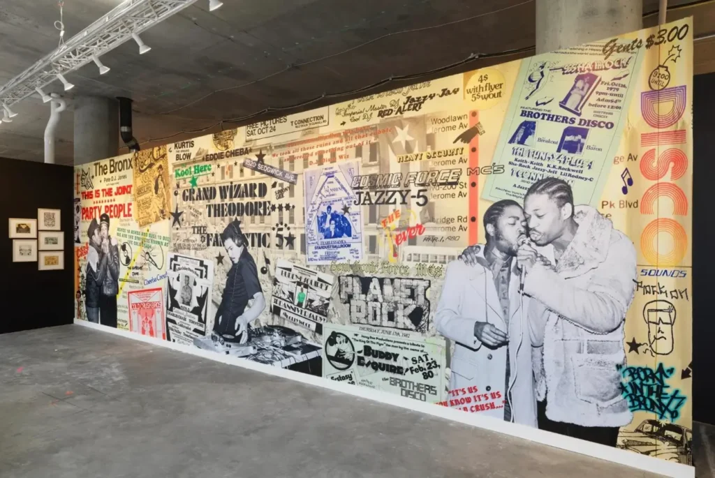 A large wall mural in a gallery space featuring a collage of early hip-hop party flyers, typography, graffiti elements, and black-and-white photographs of DJs and MCs from the Bronx. The artwork blends vintage handbills, performance announcements, and historic imagery to celebrate the origins of hip-hop culture