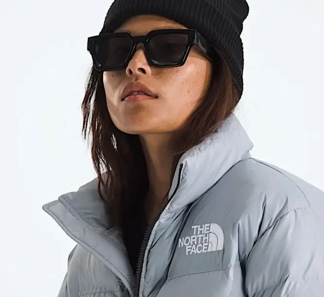 A woman wearing a light gray The North Face puffer jacket is shown in a close-up portrait. She has on oversized black rectangular sunglasses and a black knit beanie. The jacket features The North Face logo on the chest, and the high collar is zipped up. The background is a clean, soft white, highlighting her winter streetwear look