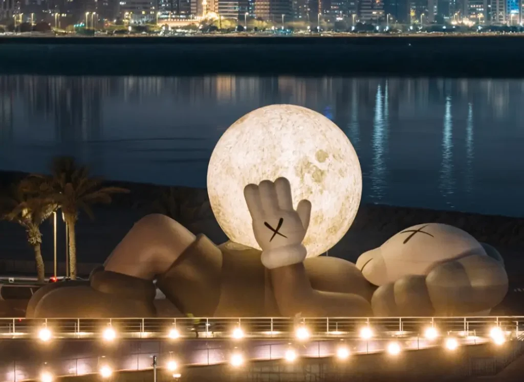 A monumental KAWS Companion sculpture lies reclined along the Abu Dhabi waterfront at night, holding a large glowing moon in its raised hand. City lights reflect across the calm water behind it, with palm trees and a lit promenade in the foreground