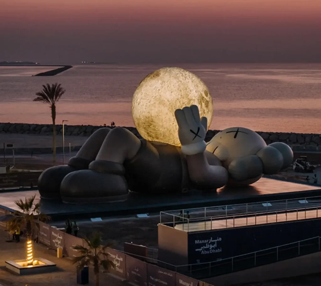 The KAWS Companion sculpture lies reclined beside the Abu Dhabi shoreline at sunset, holding a glowing moon in its raised hand. The sky fades from deep pink to purple over the calm water, with palm trees, a rock breakwater, and illuminated signage for Manar Abu Dhabi visible in the foreground