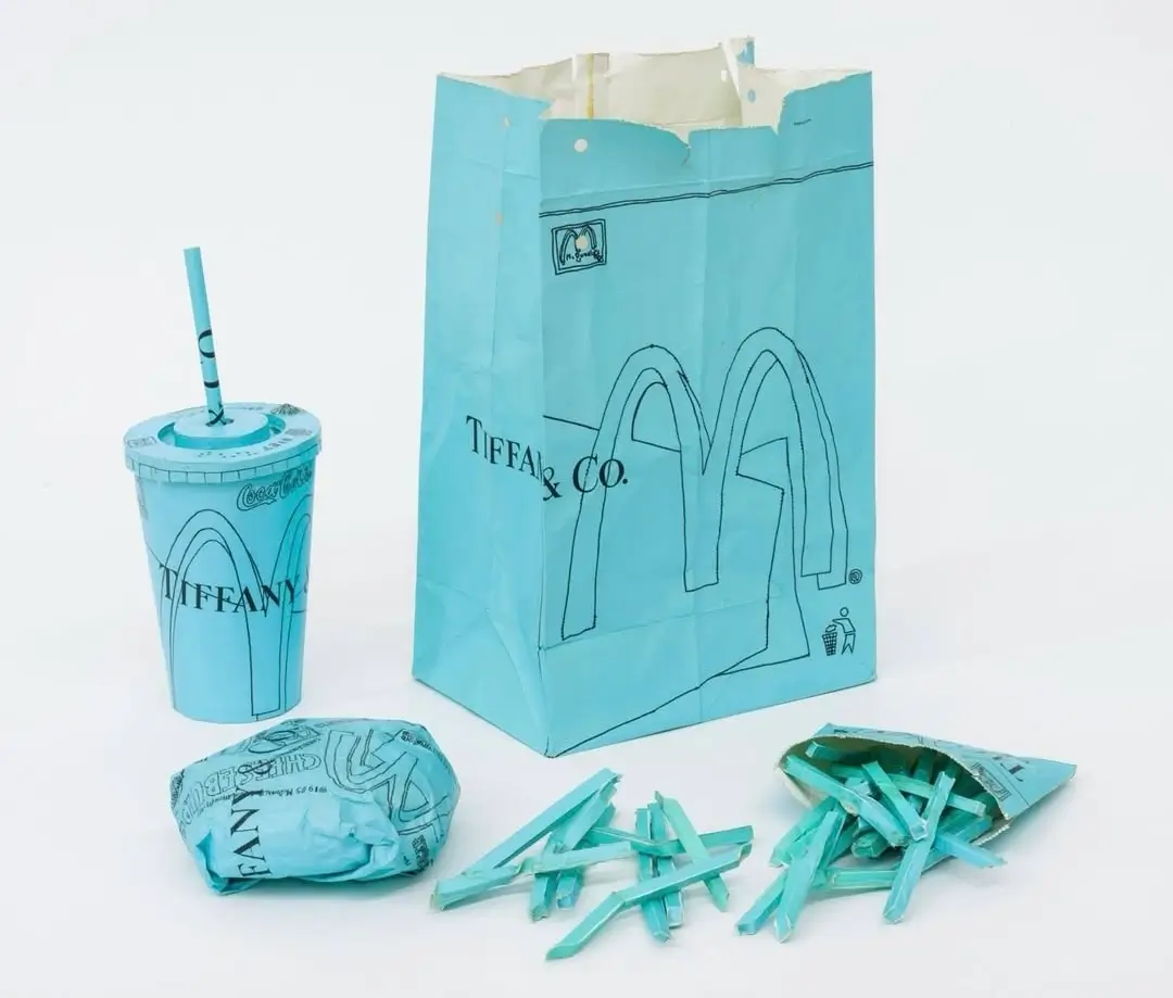 A sculptural McDonald’s meal recreated entirely in Tiffany Blue paper, featuring a tall drink cup with straw, a folded burger wrapper, scattered paper fries spilling from a small fry packet, and a crinkled takeout bag. All elements are hand-drawn with black linework, blending the Tiffany & Co. logo with McDonald’s arches, set against a clean white background