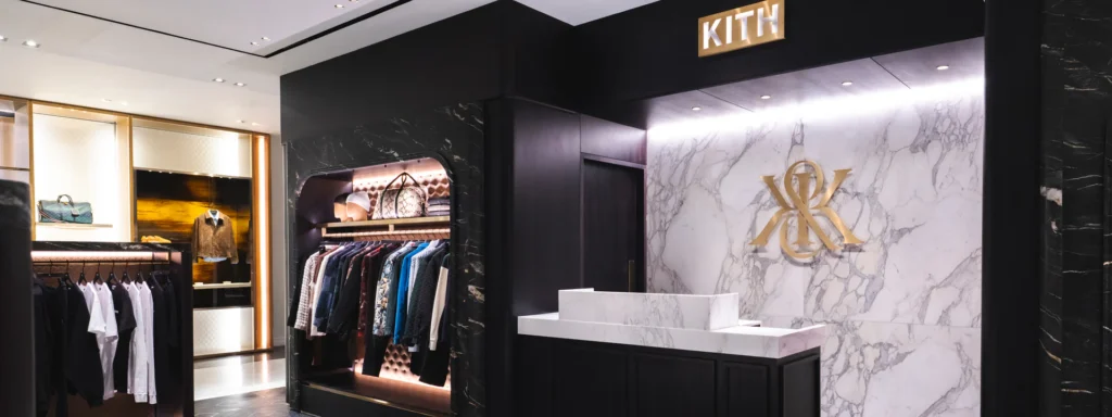 nterior of the Kith London flagship showing a marble-clad reception desk, gold Kith monogram on veined stone, and adjacent apparel displays framed in dark wood and soft lighting