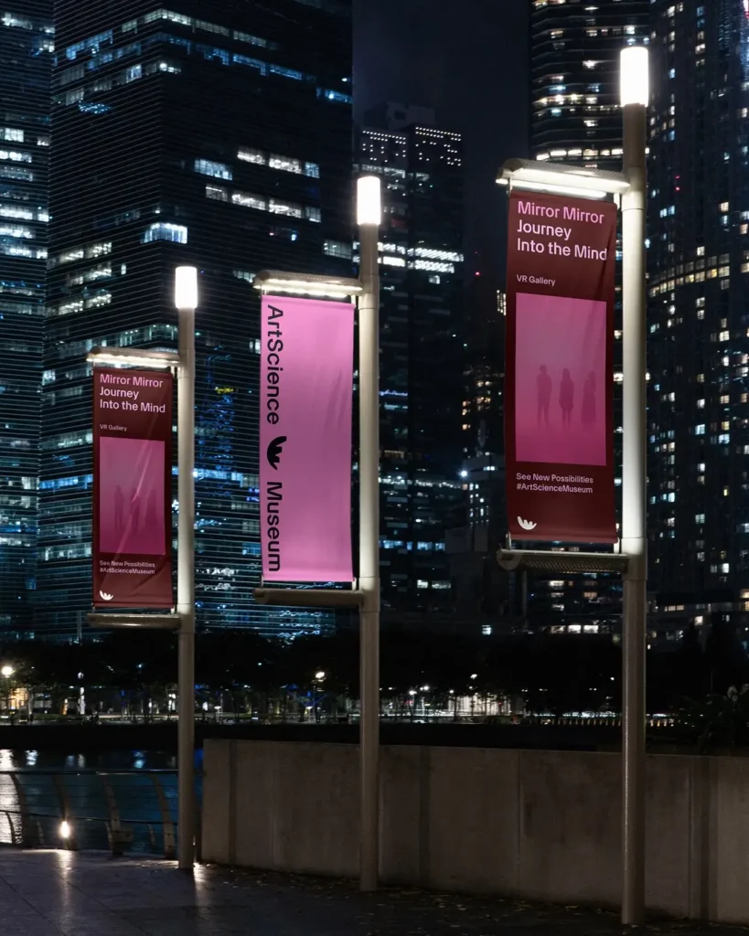 Nighttime view outside Singapore’s ArtScience Museum, featuring illuminated banners promoting the exhibition ‘Mirror Mirror: Journey Into the Mind’ at the VR Gallery. The magenta and burgundy banners stand against the Marina Bay skyline, reflecting the museum’s contemporary identity amid city lights