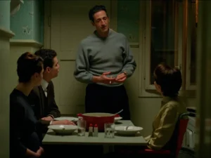 Adrien Brody, wearing the signature grey Brutalist Sweater, stands at a small dinner table in a dimly lit, vintage apartment kitchen, speaking to three seated characters as warm yellow light falls across the room and highlights the mid-century set design