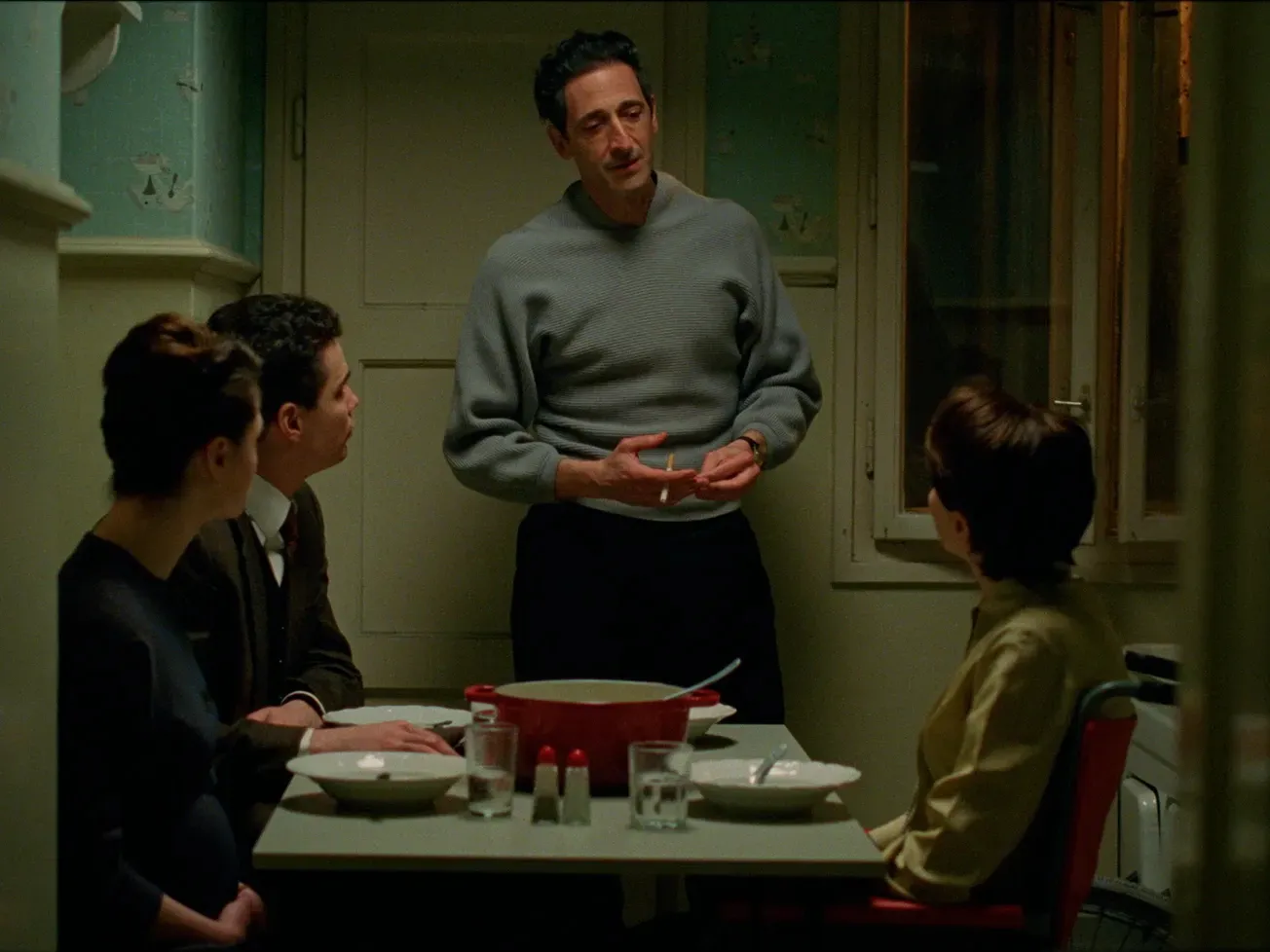 Adrien Brody, wearing the signature grey Brutalist Sweater, stands at a small dinner table in a dimly lit, vintage apartment kitchen, speaking to three seated characters as warm yellow light falls across the room and highlights the mid-century set design