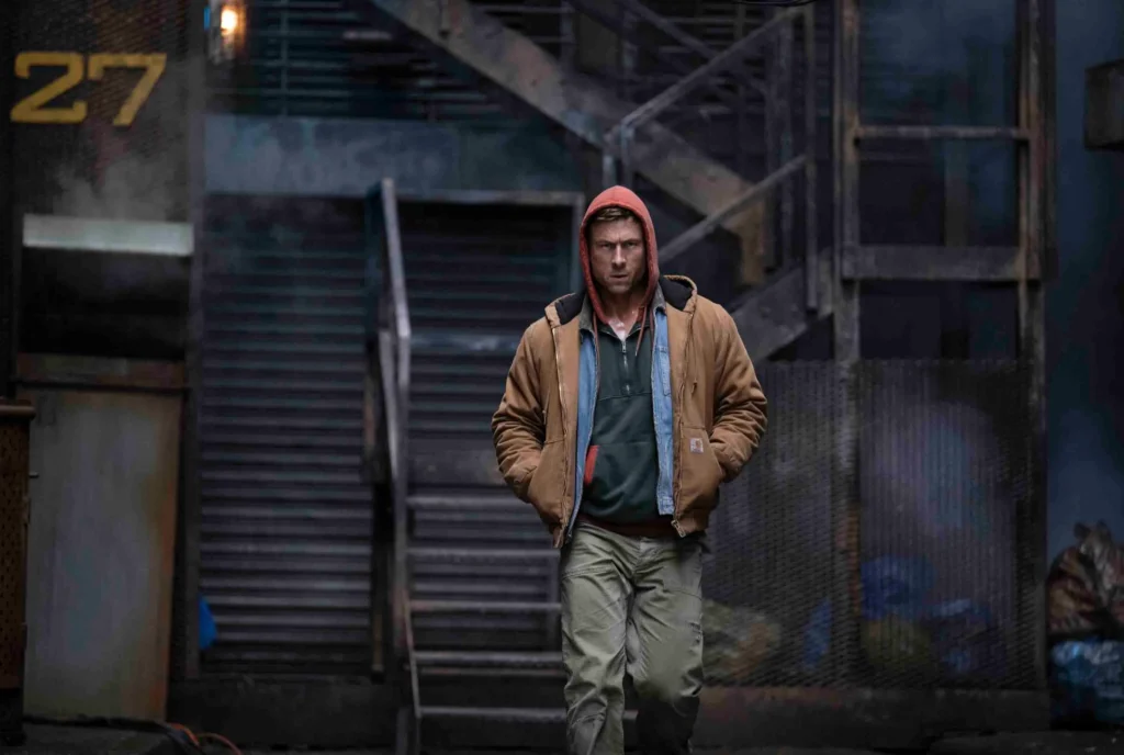Glen Powell as Ben Richards walks through a dimly lit industrial alley in The Running Man (2025), wearing layered workwear—a brown Carhartt-style jacket, red hood, and green sweatshirt—capturing the film’s gritty, blue-collar dystopian tone